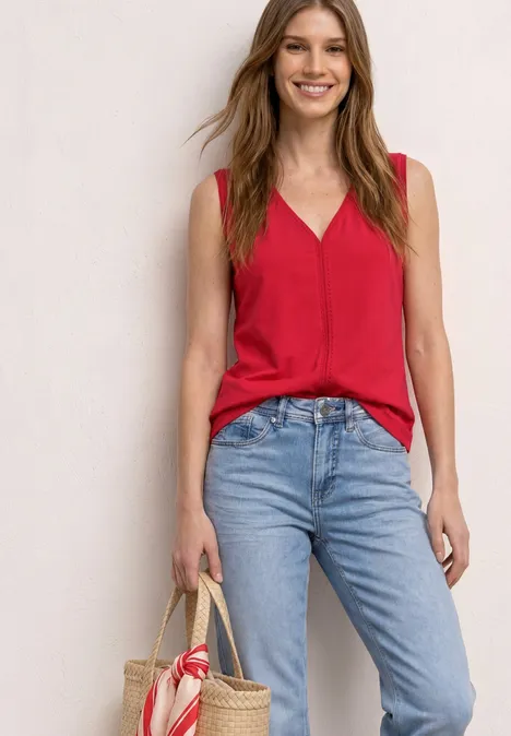 Vrouw in rood topje en blauwe jeans met strooien tas voor een egale achtergrond.