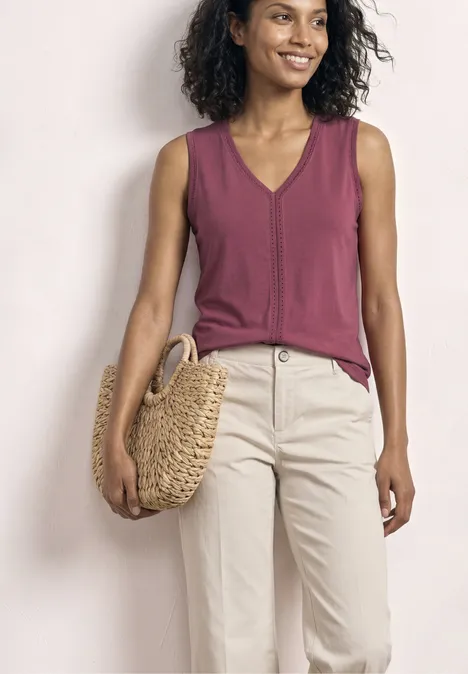 Une femme porte un débardeur violet à encolure V, un pantalon clair et tient un sac rond en paille.