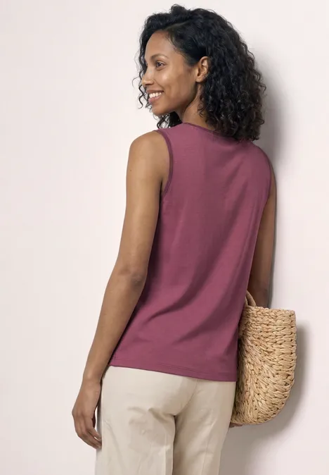 Une femme aux cheveux bouclés porte un débardeur rose et un sac tressé à l’épaule.