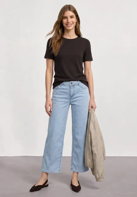 Frau in schwarzem Shirt und hellblauen Jeans hält Jacke, vor weißem Hintergrund.