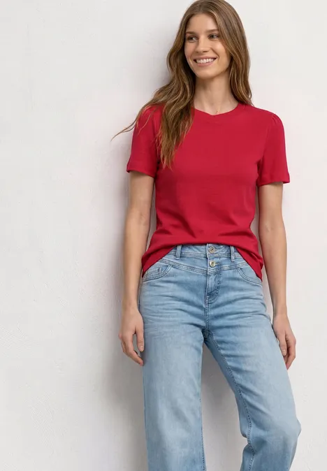 Frau in rotem T-Shirt und blauen Jeans steht vor hellem Hintergrund.