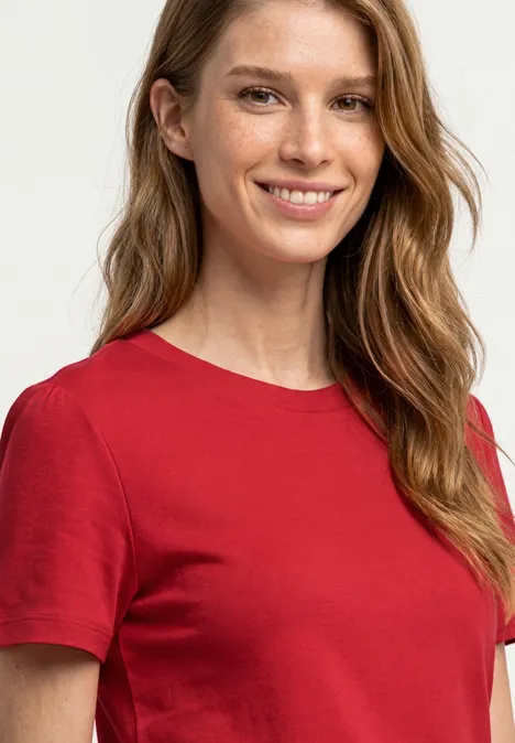 Eine Frau mit langen blonden Haaren trägt ein rotes T-Shirt vor hellem Hintergrund.