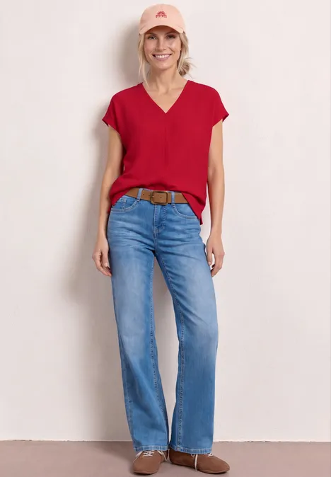 Vrouw in rood topje, blauwe jeans, bruine riem en enkellaarzen voor lichte achtergrond.