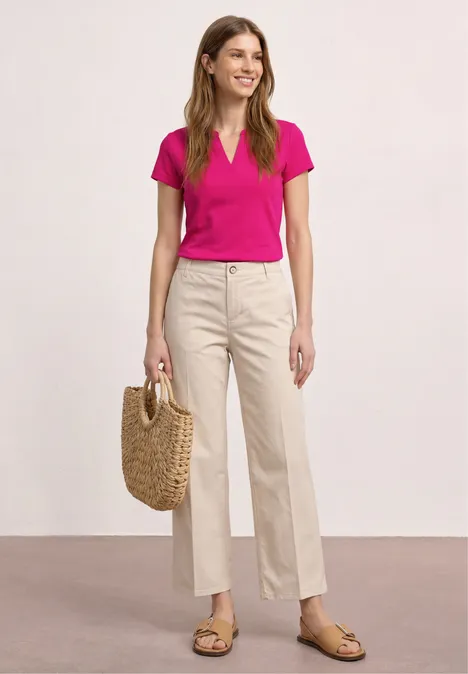 Frau in pinkem T-Shirt, beigen weiten Hosen, Strohtasche und Sandalen vor neutralem Hintergrund.