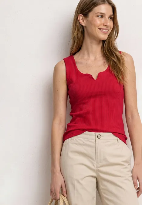 Femme debout, porte un top rouge sans manches et un pantalon beige.
