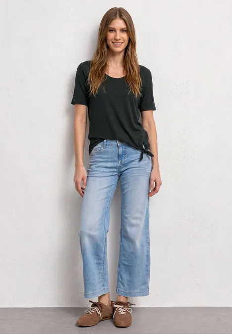 Frau steht vor weißer Wand in schwarzem T-Shirt, weitem blauen Jeans und braunen Schuhen.