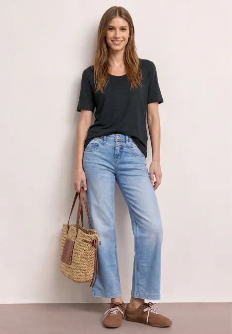 Frau in schwarzem T-Shirt, hellen, weiten Jeans, braunen Sandalen und Korbtasche vor neutralem Hintergrund.