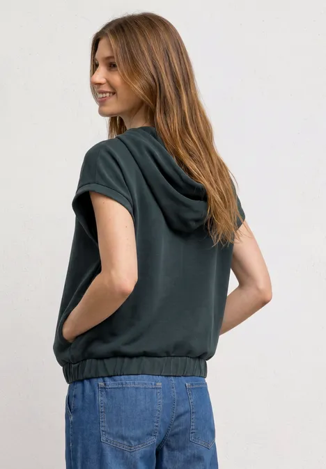 Eine Frau in einem ärmellosen Kapuzenpullunder steht mit den Händen in den Taschen vor hellem Hintergrund.