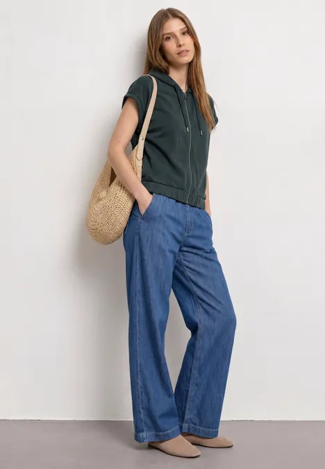 Frau in schwarzem Kapuzenshirt, weiten blauen Jeans, braunen Schuhen und Strohtasche.