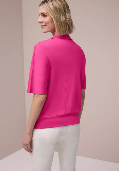 Eine Frau steht von hinten vor hellem Hintergrund, trägt ein pinkes Kurzarmoberteil und weiße Hosen.