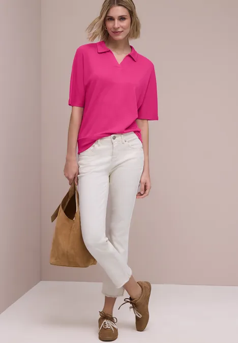Eine Person vor neutralem Hintergrund trägt ein pinkes Poloshirt, weiße Jeans, braune Schuhe und hält eine Tasche.