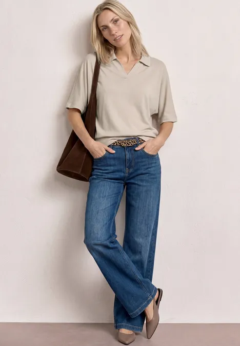 Person mit grauem Poloshirt, blauen Jeans, brauner Schultertasche und Loafers vor neutralem Hintergrund.