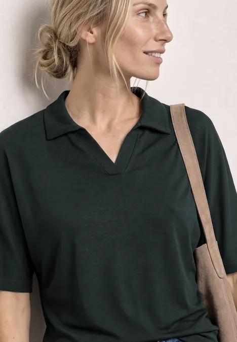 Vrouw in een donker poloshirt, haar in een losse knot, draagt een stoffen tas over haar schouder.