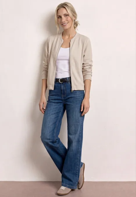 Frau in weißem T-Shirt, beiger Jacke, weiten Jeans und Loafers vor hellem Hintergrund.