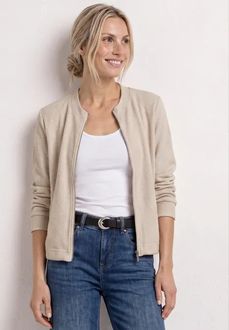 Eine Frau trägt eine beigefarbene Strickjacke, ein weißes Top und blaue Jeans mit Gürtel.