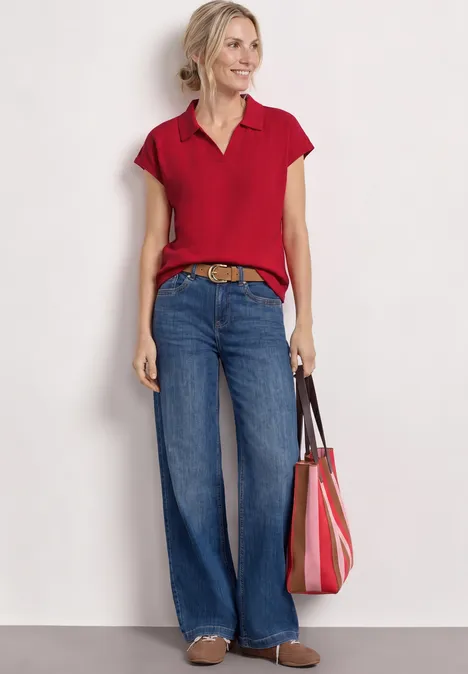 Frau mit rotem Polo, weiten Jeans und roter Handtasche vor hellem Hintergrund.