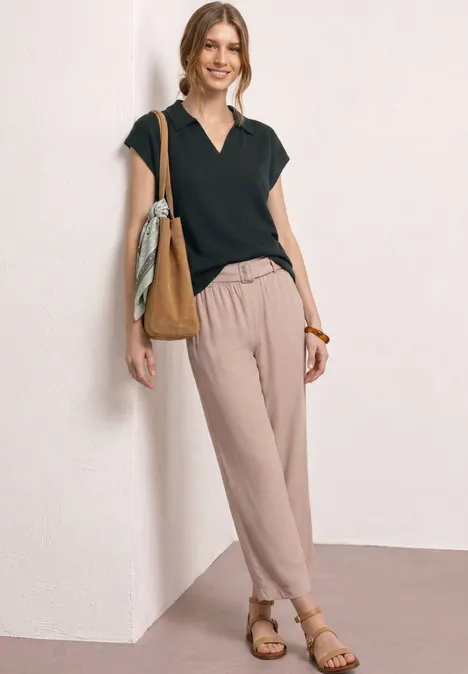 Frau lehnt lässig an Wand, trägt schwarzes V-Shirt, helle Hose, Sandalen und Umhängetasche.