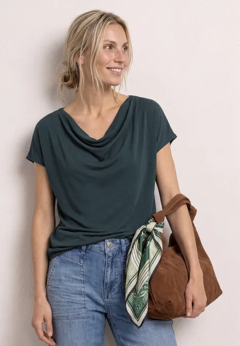Eine Frau trägt ein dunkelgrünes Wasserfall-Shirt, Jeans und hält eine braune Tasche mit grünem Halstuch.