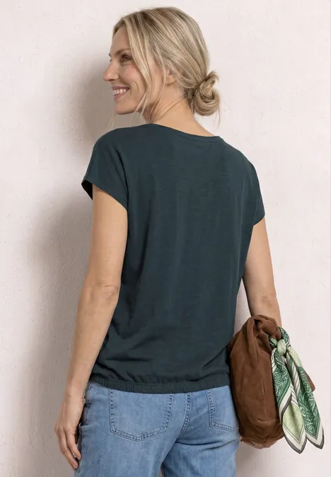 Frau von hinten mit dunklem T-Shirt und Jeans, hält braune Tasche mit grün gemustertem Tuch.