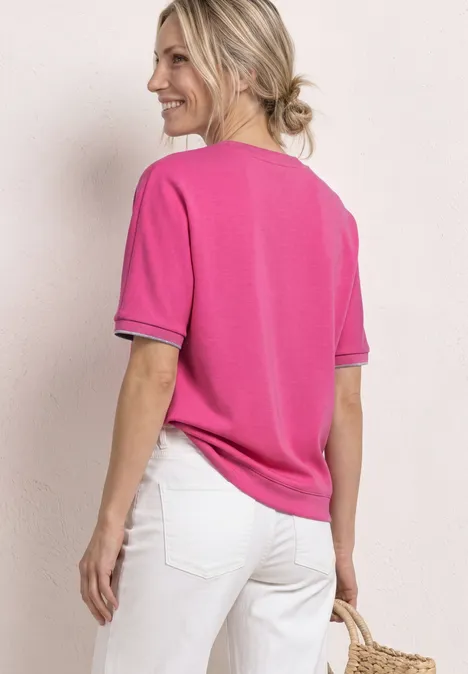 Eine Frau von hinten in pinkem Kurzarmshirt mit schwarzem Saum, weißen Hosen und geflochtener Korbtasche.