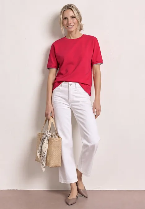 Frau in rotem T-Shirt, weißer Culotte, grauen Pumps, hält eine geflochtene Korbtasche mit Tuch.