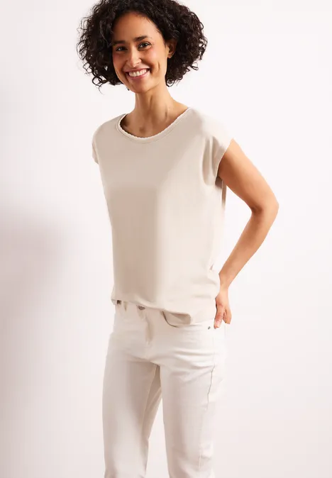Frau in weißem T-Shirt und weißen Hosen steht vor hellem Hintergrund.
