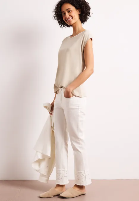 Frau trägt beiges Top und weiße Hose, hält leichte Jacke in der Hand vor weißem Hintergrund.