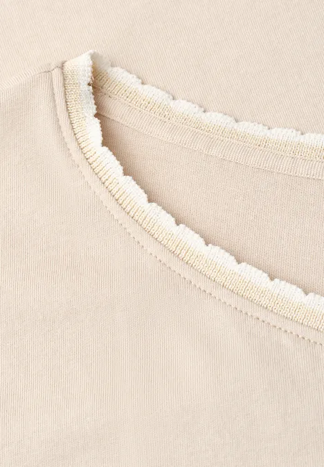 Nahaufnahme eines beigefarbenen Kleidungsstücks mit feinem, gewelltem Spitzenbesatz am Halsausschnitt.