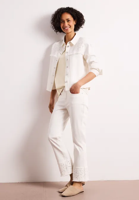 Frau mit lockigen Haaren trägt weiße Jeansjacke und Hose, beige flache Schuhe, lehnt lässig an heller Wand.