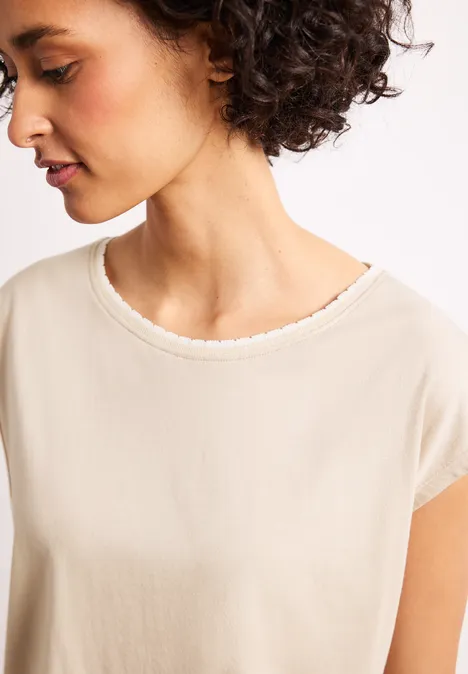 Person mit lockigem dunklem Haar trägt cremefarbenes T-Shirt mit Rundhalsausschnitt.