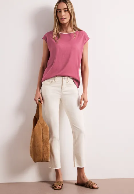 Een vrouw in een roze top, witte cropped jeans, sandalen en bruine tas voor een lichte achtergrond.