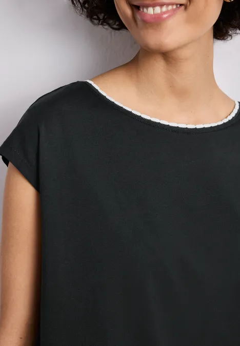 Een glimlachende vrouw draagt een eenvoudig zwart shirt met witte ronde hals, haar krullen omlijsten het gezicht.