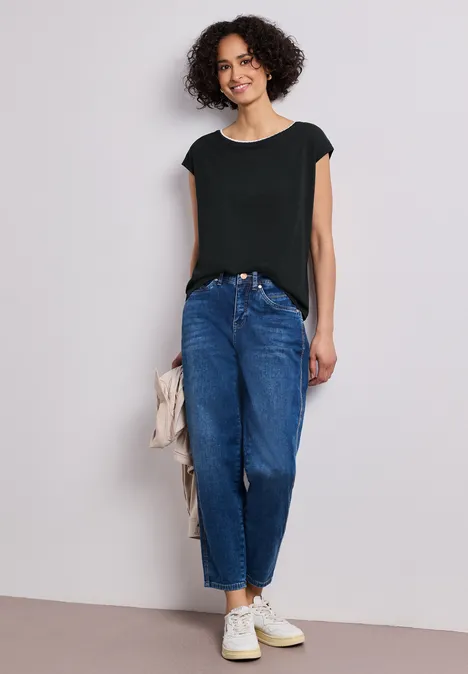 Een vrouw met krullend haar draagt een zwart topje, blauwe jeans en witte sneakers, met een beige jasje in haar hand.