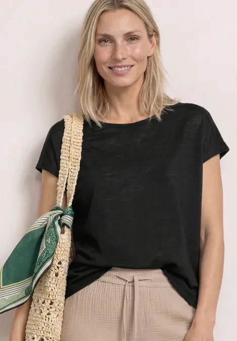 Vrouw in zwarte t-shirt, beige broek en gevlochten schoudertas met een groen sjaaltje.
