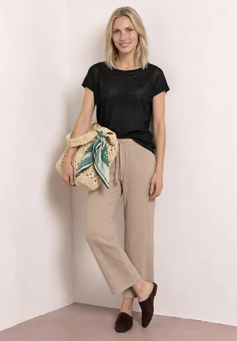 Vrouw in zwart t-shirt, beige broek en bruine loafers voor een witte muur, met een gevlochten tas.