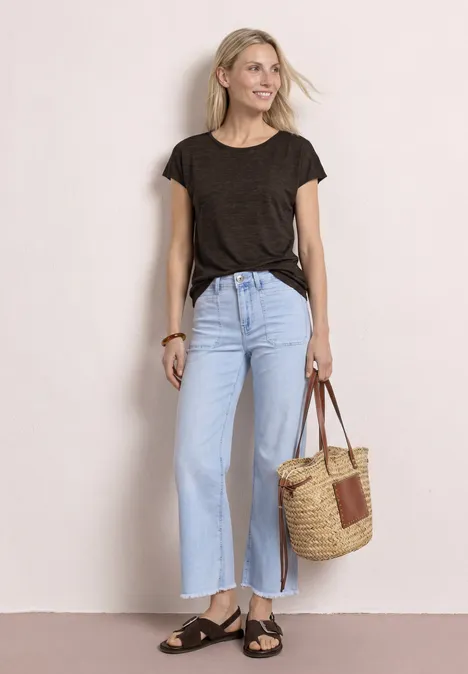 Vrouw in zwart T-shirt, lichtblauwe wijde jeans, sandalen en rieten tas voor een witte achtergrond.