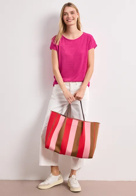 Iemand staat voor een lichte muur, draagt een roze shirt, witte broek, witte sneakers en houdt een gestreepte shopper vast.