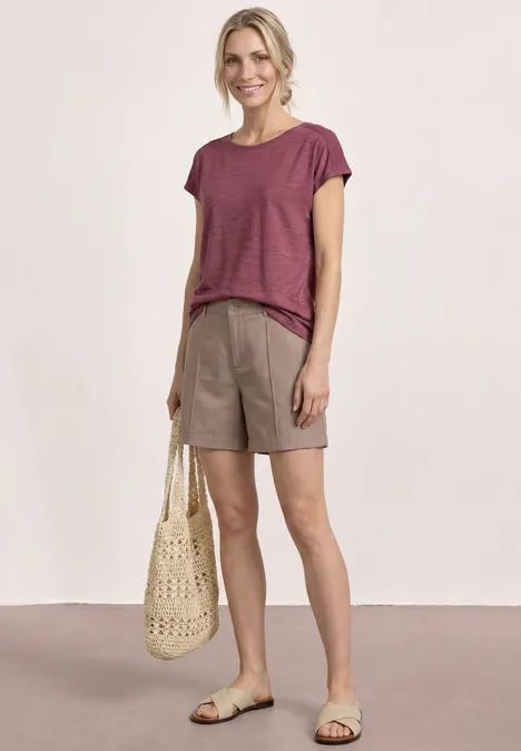 Vrouw in een relaxte zomerse look met bordeaux t-shirt, beige short, gevlochten tas en sandalen.