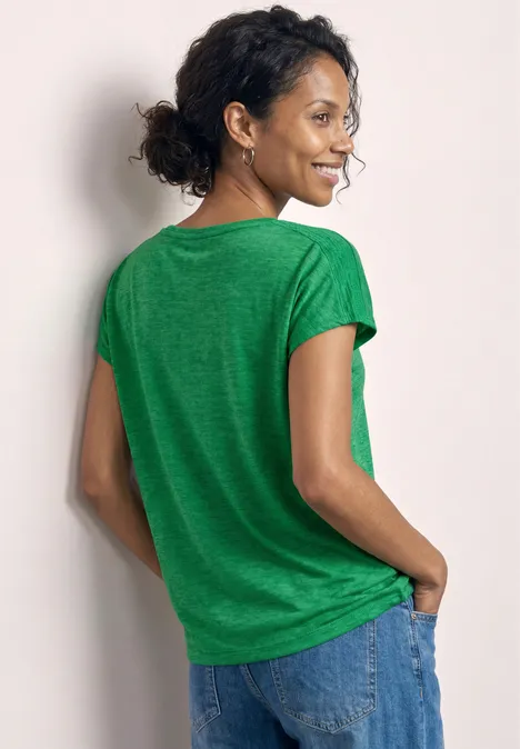 Een persoon met de rug naar de camera draagt een groen t-shirt en jeans, handen in de zakken.