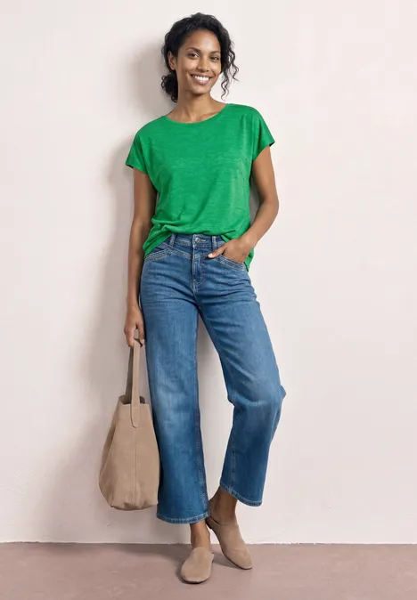 Vrouw in groen T-shirt, wijde spijkerbroek, beige enkellaarzen en tas, staat nonchalant met één hand in haar zak.