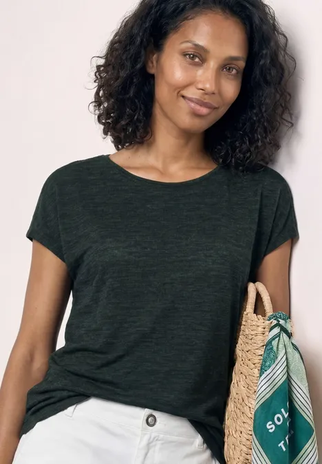 Vrouw met krullend haar, donkergroen T-shirt, witte broek, houdt een mand met een groen sjaaltje vast.