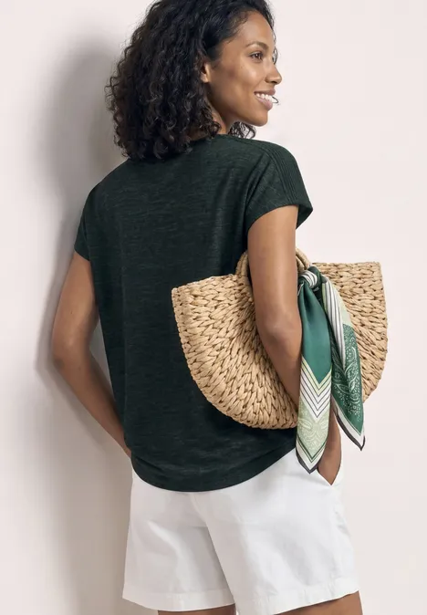 Vrouw van achteren in donkergroen shirt en witte shorts, draagt strooien tas met groene sjaal