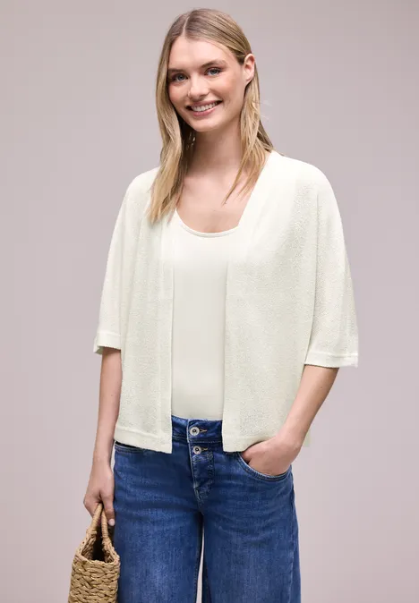 Vrouw draagt crème kort vest over top, blauwe jeans en heeft een strooien tas in de hand.