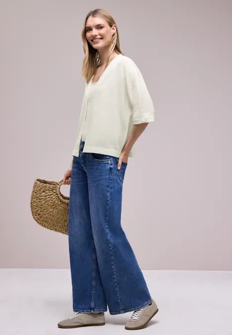 Vrouw in wijde blauwe jeans en witte blouse, met sneakers en gevlochten strooien tas.