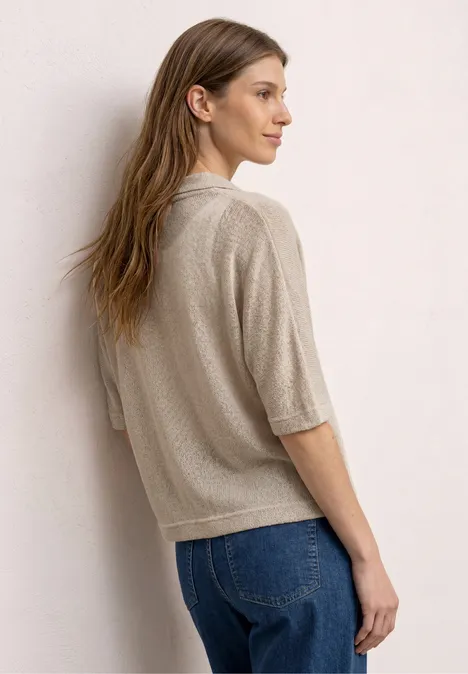 Vrouw met rug naar camera, draagt beige trui en blauwe jeans, leunend tegen een lichte muur.
