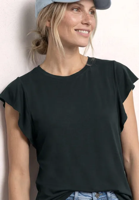 Frau in schwarzem Top mit Rüschenärmeln, Jeans und Baseballcap vor hellem Hintergrund.