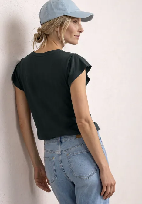 Frau von hinten in schwarzem T-Shirt, Jeans und Cap vor weißer Wand