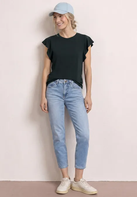 Frau in schwarzem Shirt, hellblauen Jeans, weißen Sneakern und Cap vor neutralem Hintergrund.