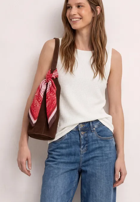 Frau in weißem ärmellosem Oberteil, blauen Jeans, trägt braune Tasche mit rotem Halstuch.