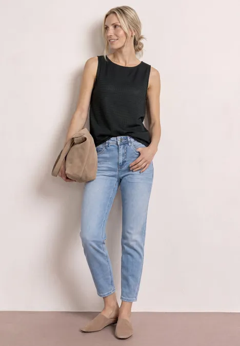 Vrouw in zwarte mouwloze top, lichtblauwe jeans, beige loafers en tas tegen een neutrale achtergrond.